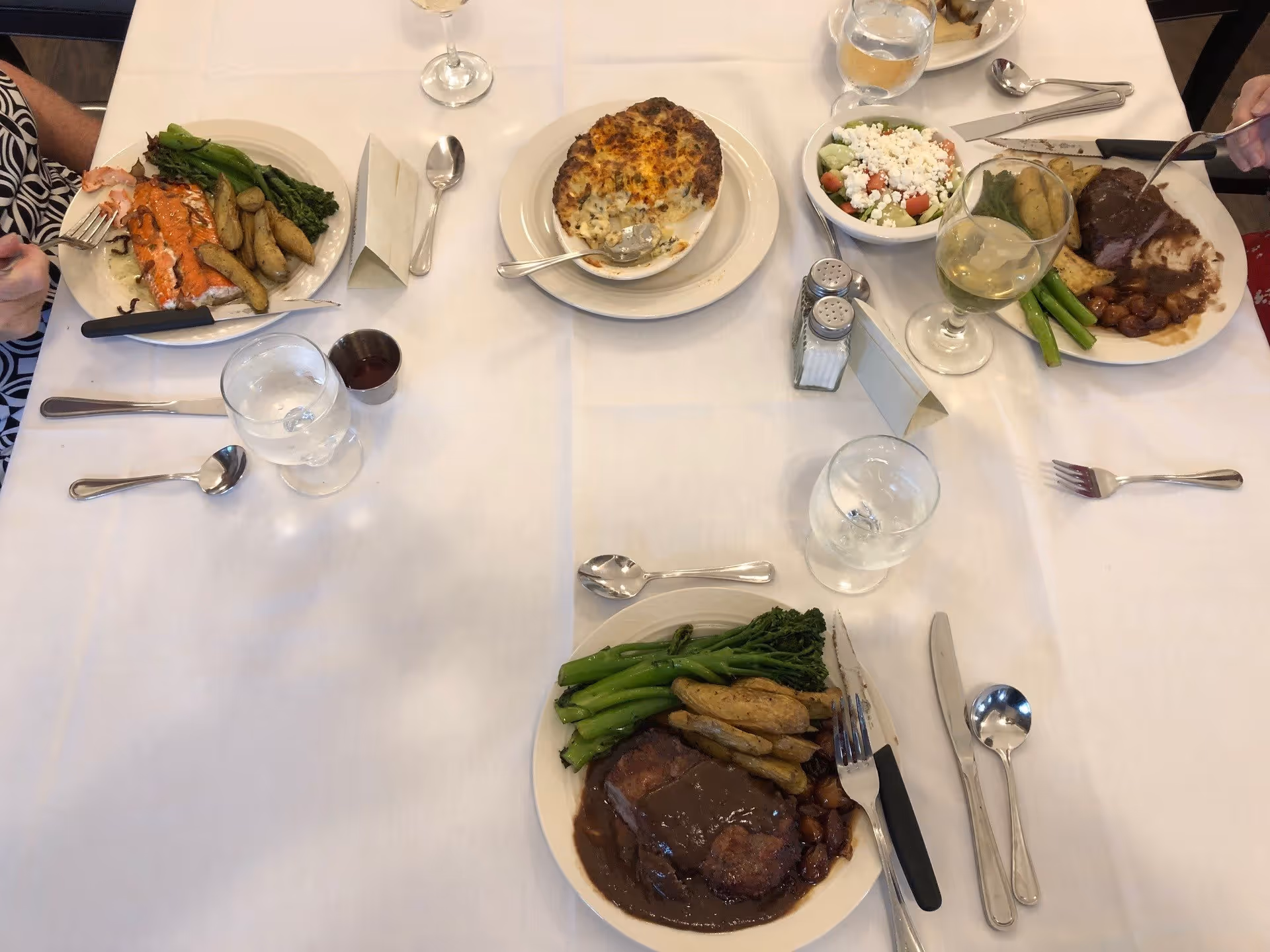 A dining table set with three plates of food including meat with gravy, roasted potatoes, and green vegetables. There is also a bowl of salad with feta cheese and a dish of baked casserole. The table is covered with a white tablecloth and has glasses of water and wine, silverware, and salt and pepper shakers.