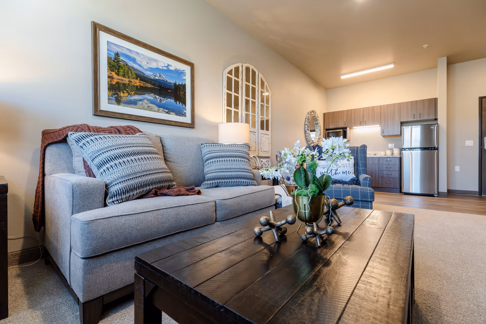 A cozy senior living room with a gray sofa adorned with patterned pillows and a brown throw blanket. A dark wooden coffee table with decorative items and a potted plant sits in front of the sofa. In the background, there is a kitchen area with wooden cabinets, a stainless steel refrigerator, and a microwave. The walls are decorated with a framed landscape photograph, a rustic window frame, and a round mirror. The room has warm lighting and a carpeted floor.