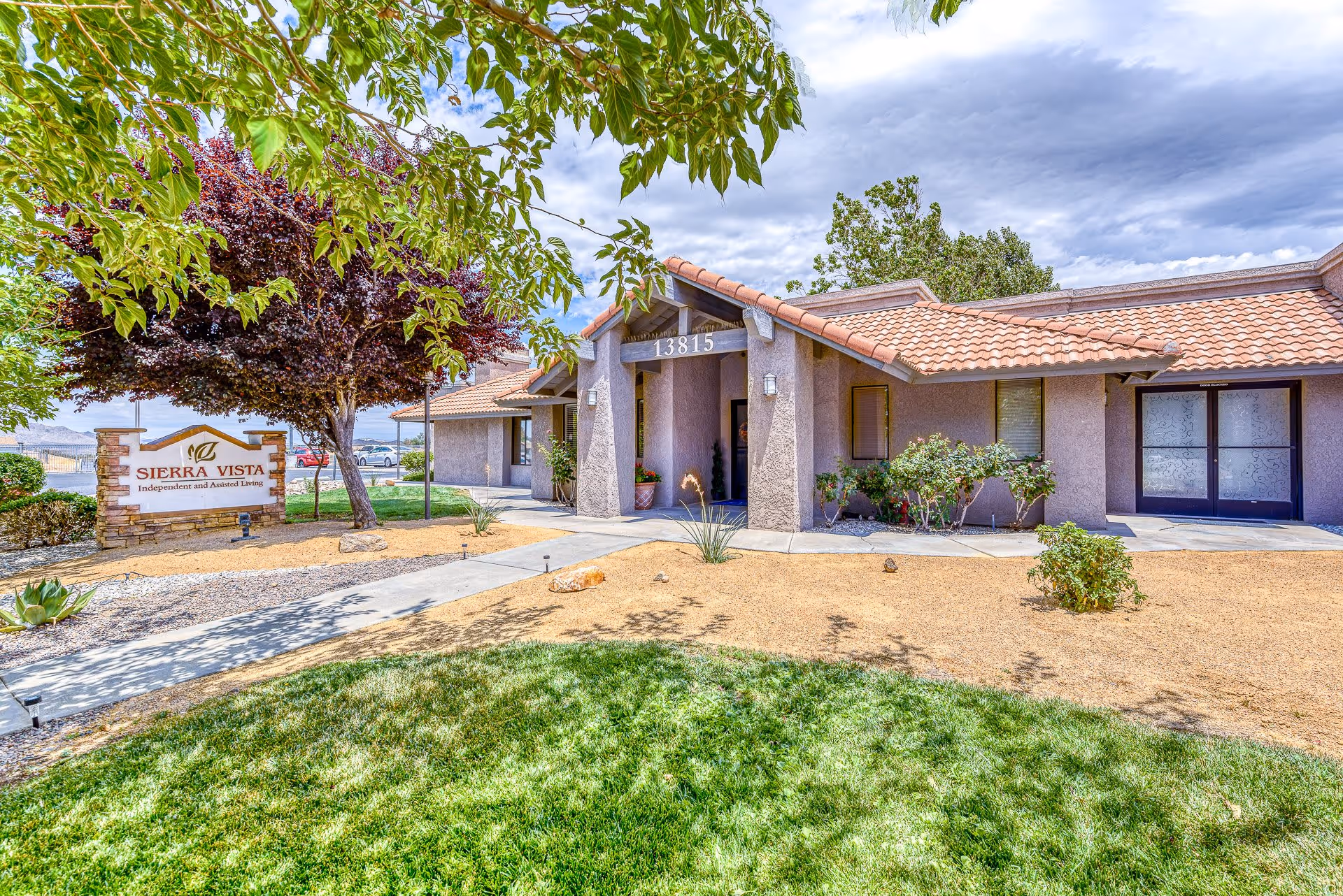 Exterior view of Sierra Vista Independent and Assisted Living facility showing a single-story building with a tiled roof, surrounded by landscaped grounds with green grass, trees, shrubs, and a clear pathway leading to the entrance. A sign with the facility name is visible on the left side near the sidewalk.