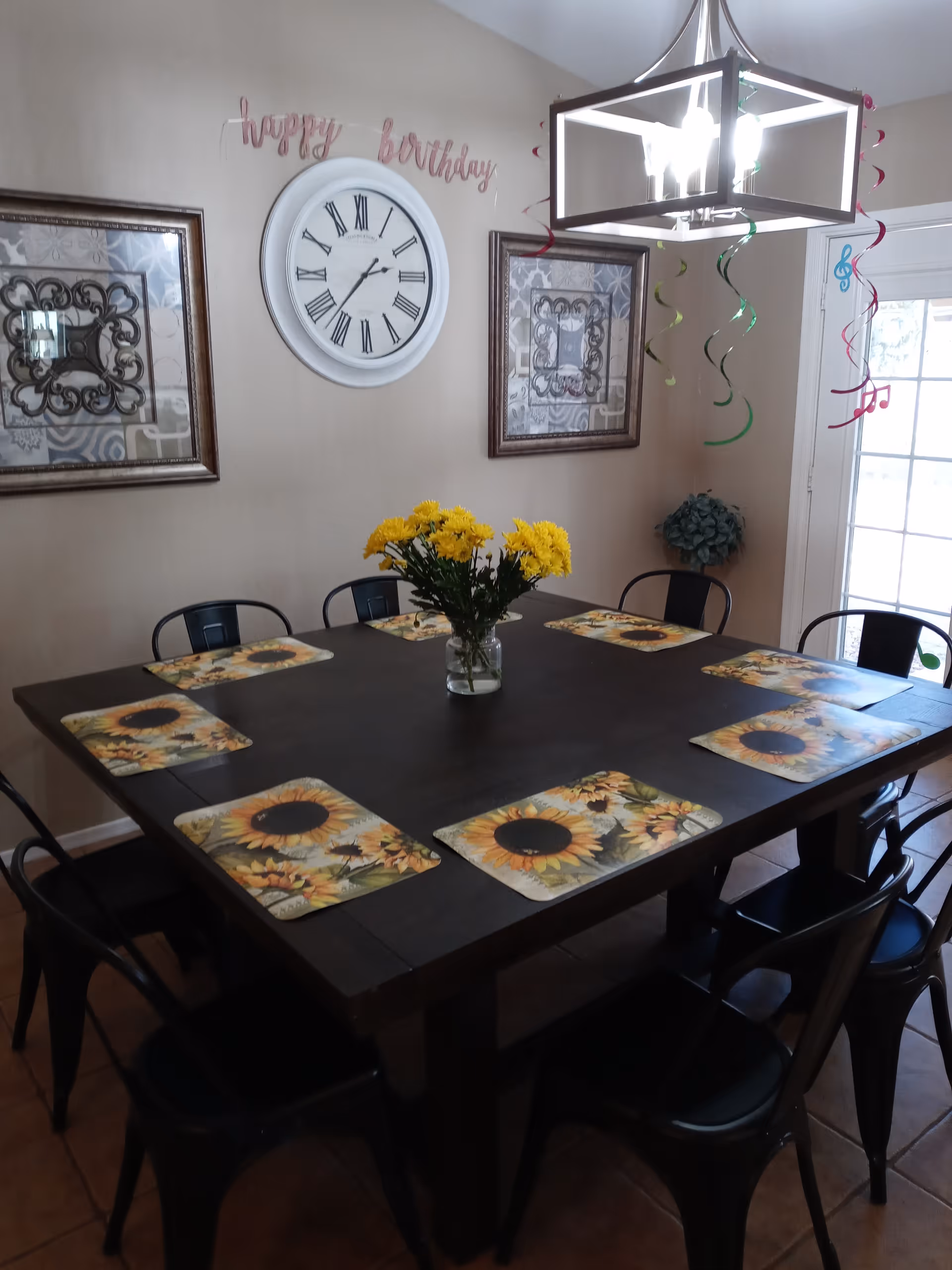 A dining room with a dark wooden square table set with eight sunflower-themed placemats and black metal chairs. A vase with yellow flowers is centered on the table. The walls are beige with two framed decorative artworks and a large white clock showing the time as 8:20. Above the clock, there is a pink 'happy birthday' banner. Colorful hanging streamers and musical note decorations are suspended from a modern square chandelier. A glass door is visible on the right side.