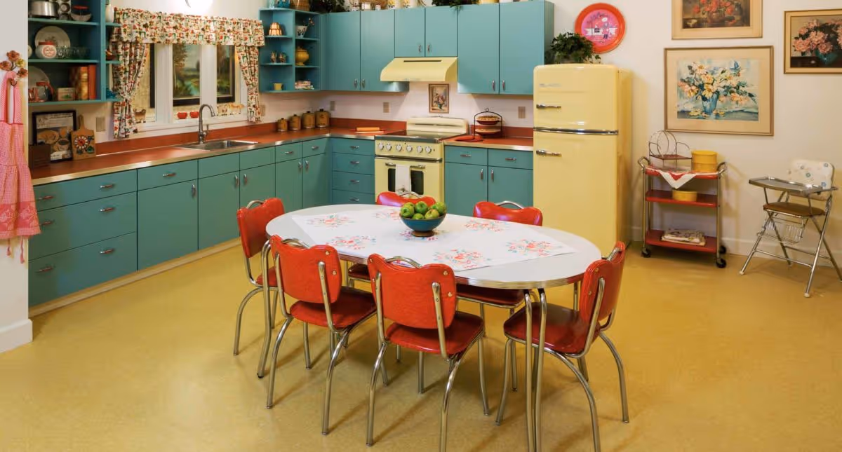 A retro-style kitchen with teal cabinets, a yellow refrigerator, and a matching yellow stove. There is a white oval table with a floral tablecloth and six red chairs around it. The kitchen has a window with floral curtains, various kitchen items on the counters and shelves, and framed artwork on the walls.