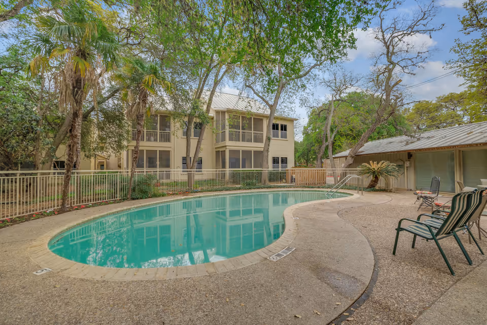 Outdoor swimming pool surrounded by a concrete deck with several lounge chairs and tables. The pool area is fenced and shaded by tall trees. In the background, there are two-story residential buildings with screened balconies and large windows.