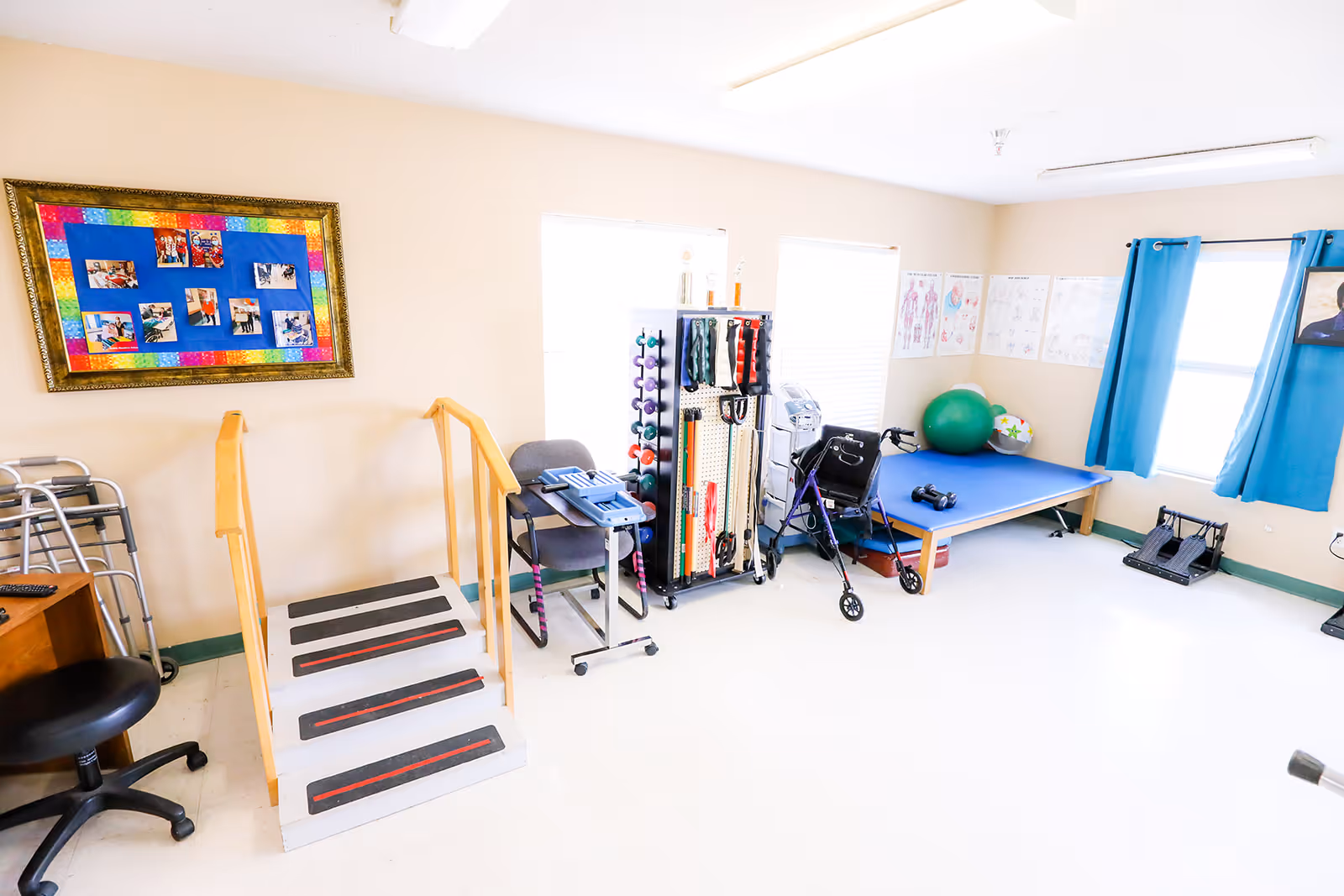 Bright rehabilitation therapy room with exercise stairs and rails, weights and resistance equipment, a balance ball, walker and treatment mat near windows with blue curtains.