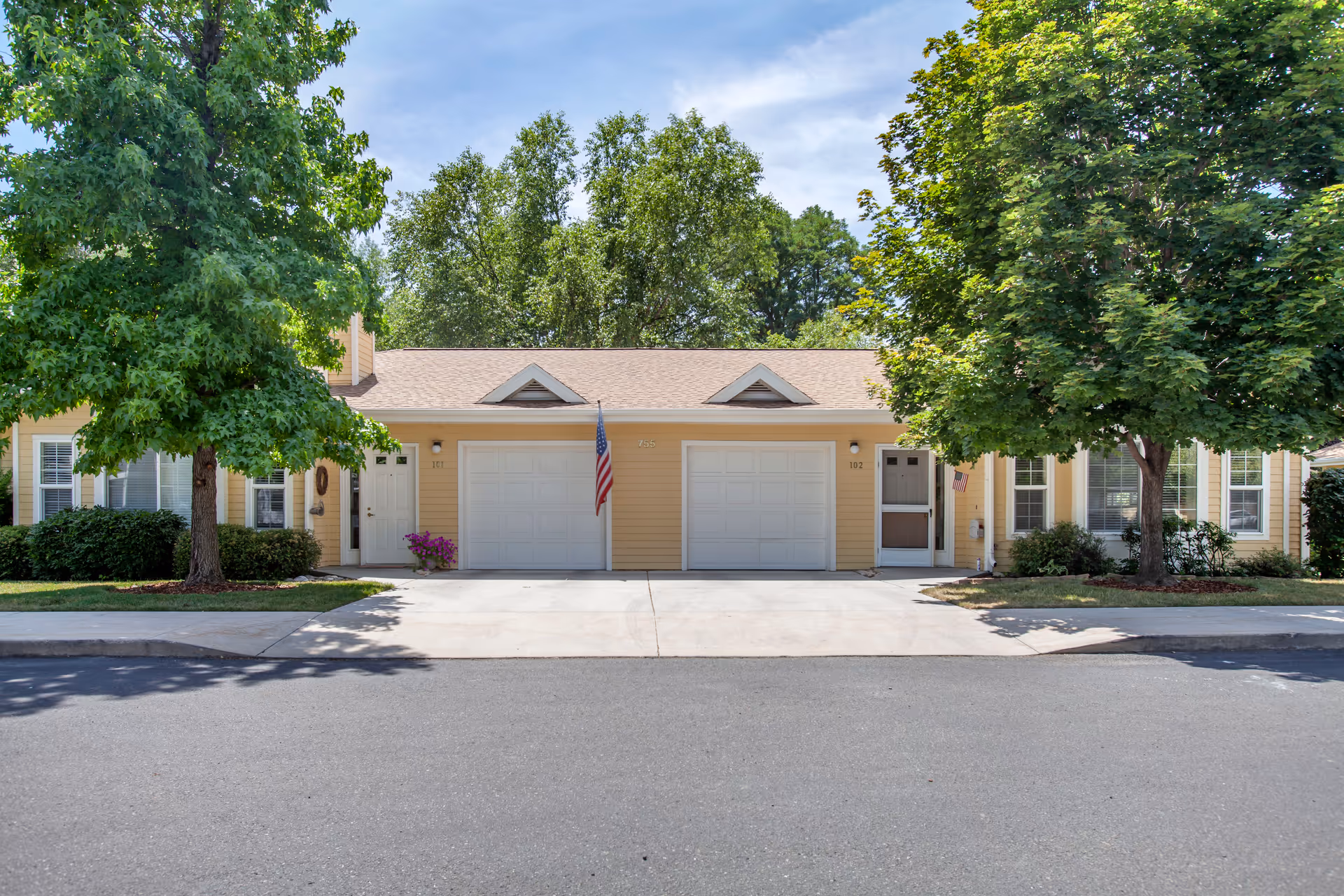 Front view of a beige senior living complex with garages, trees, and a flag.