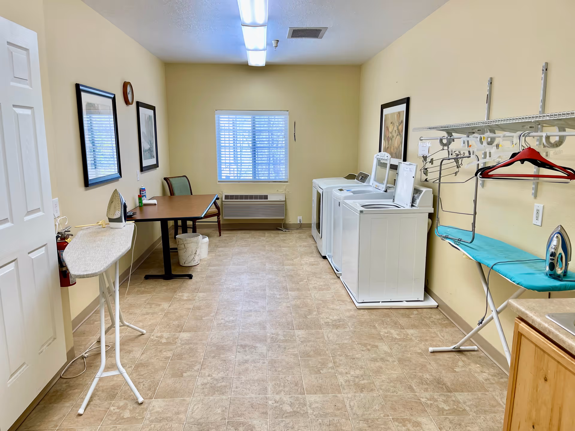 A laundry room with two washing machines and a dryer along the right wall, an ironing board with an iron, a small table with two chairs, a wall-mounted shelf with hangers, and a window with blinds at the far end. The room has beige walls and tiled flooring.