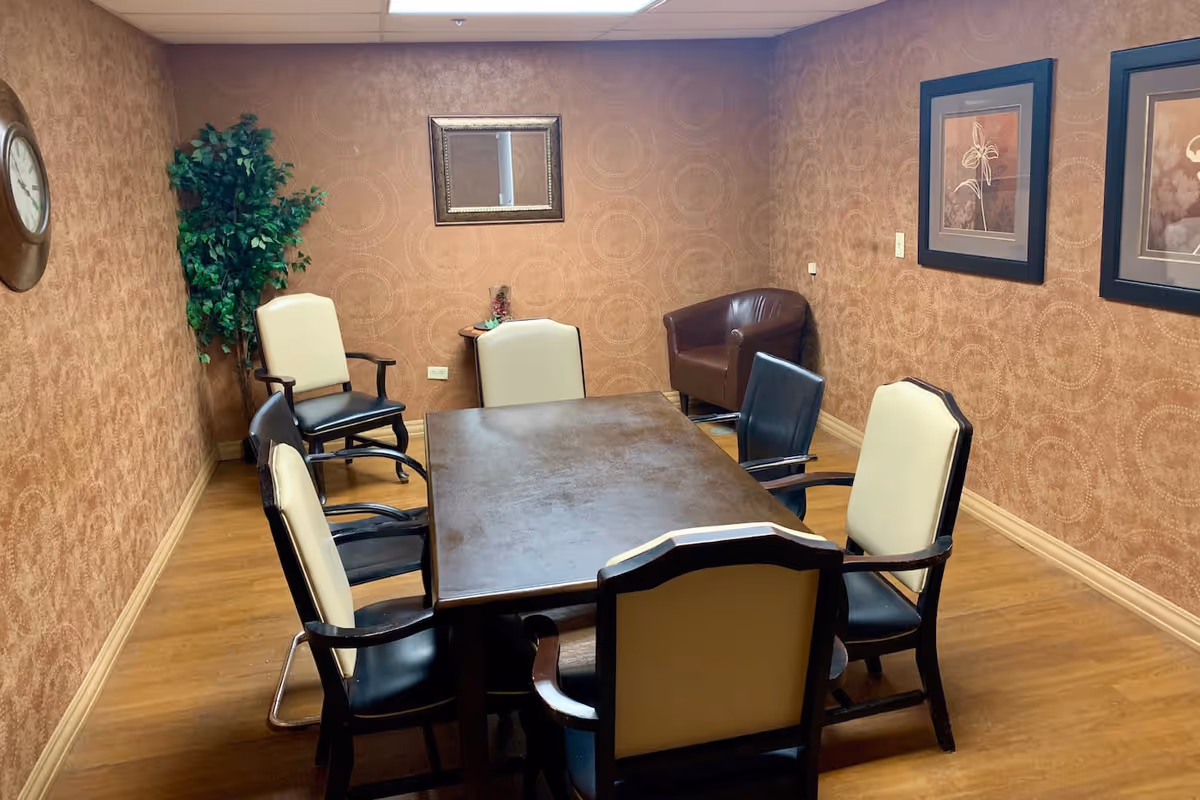 A small meeting or dining room with a rectangular wooden table surrounded by six chairs, some with cream-colored backs and others with black backs. The room has brown patterned wallpaper, wooden flooring, a wall clock, framed artwork, a small side table with decorative items, and a potted plant in the corner.