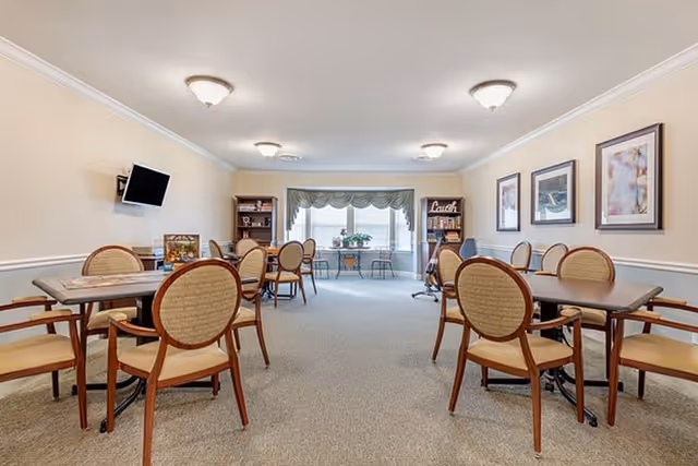 A well-lit dining room with multiple tables and chairs arranged neatly. The room features beige walls with white trim, ceiling lights, framed artwork on the walls, a wall-mounted TV, bookshelves, and a large window with a decorative valance letting in natural light.