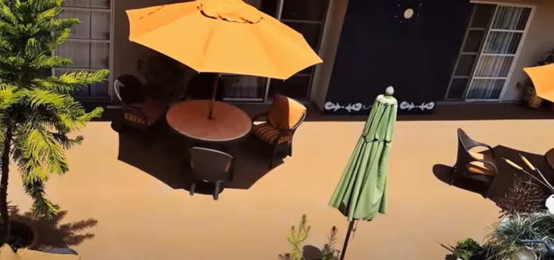 Sunlit outdoor courtyard with round tables, orange umbrellas, chairs, and potted plants in front of building doors.