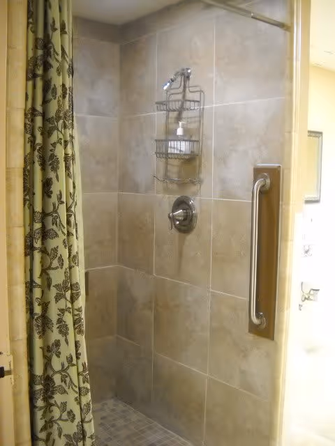 A tiled shower area with beige and light brown tiles, a metal shower handle, a wall-mounted shower caddy holding soap and shampoo, and a floral patterned shower curtain partially drawn to the side.