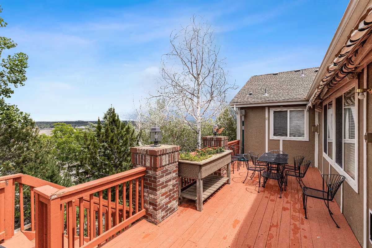 Outdoor wooden deck area with red railing and brick pillars, featuring a planter box with flowers, several black metal chairs and tables, adjacent to a building with windows and an awning, surrounded by trees under a clear blue sky.