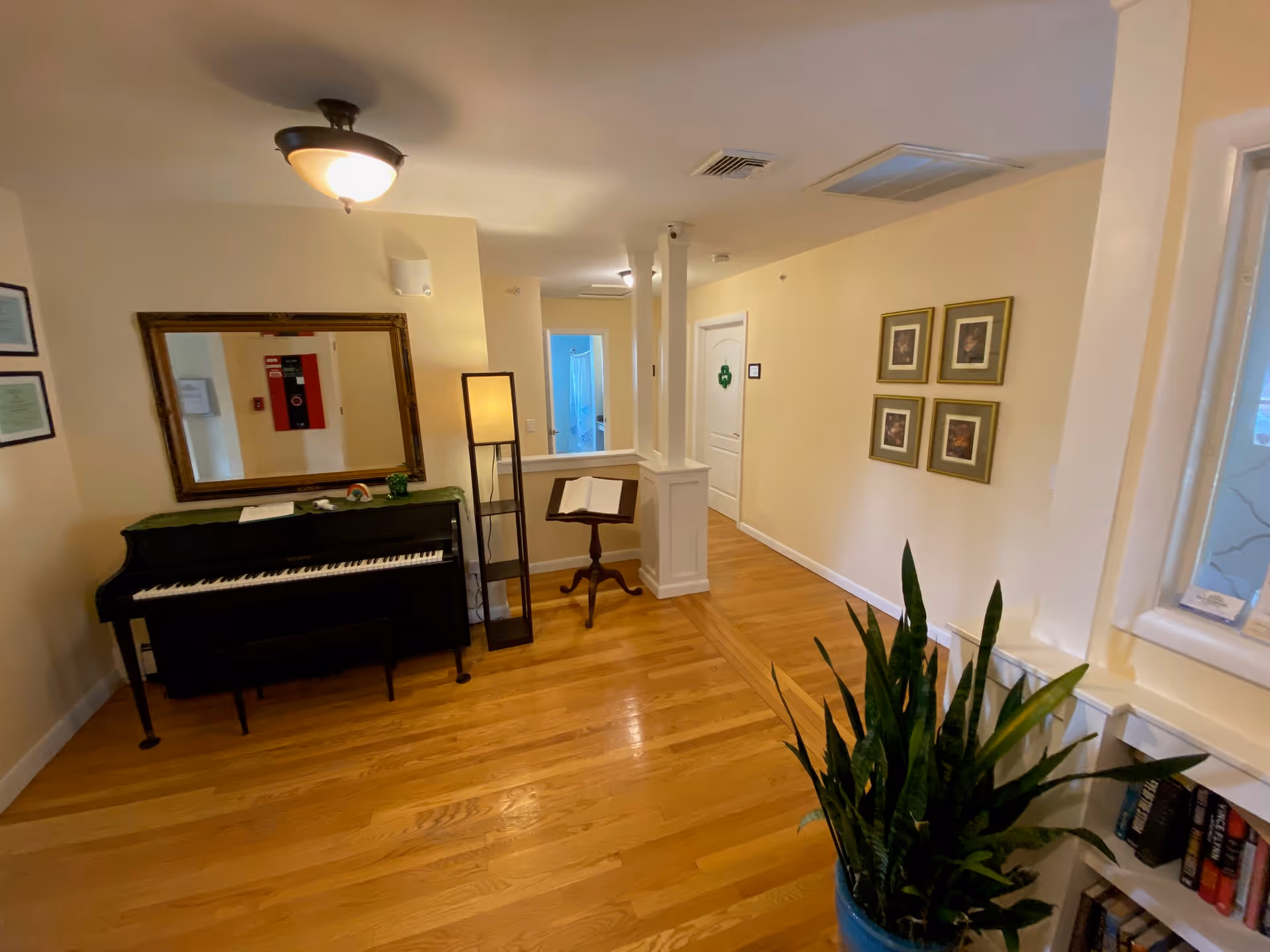Interior view of a hallway area in an assisted living facility with wooden flooring, a black upright piano with a bench, a large mirror above the piano, a tall floor lamp, a music stand with an open book, framed pictures on the wall, and a large potted plant near a window with books on the windowsill.