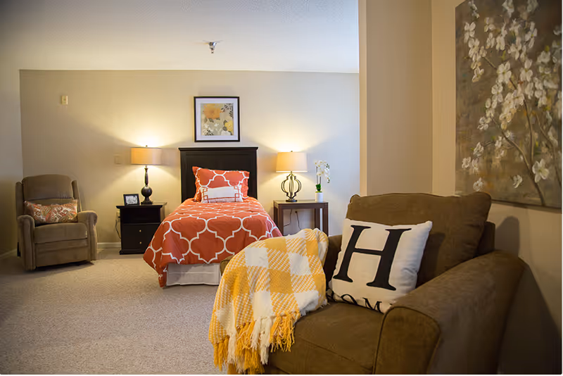Cozy senior living bedroom with a single bed, nightstands and lamps, a recliner and an armchair with a throw blanket.