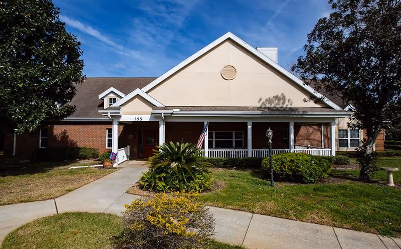 Front exterior view of a single-story brick building with a beige gable roof, a covered porch with white railings, an American flag near the entrance, and landscaped greenery including bushes and trees under a blue sky.