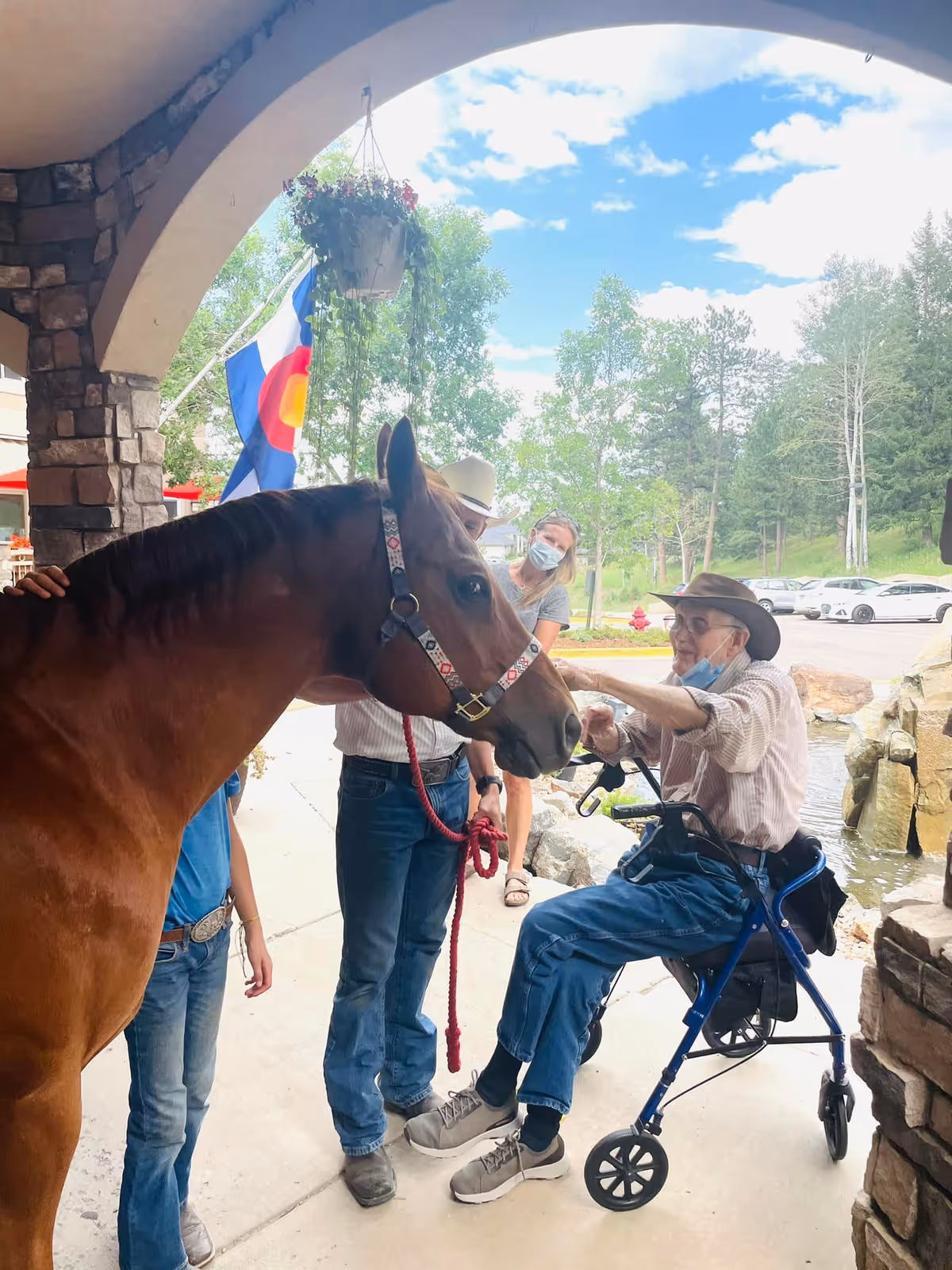 An elderly man sitting on a blue walker is petting a brown horse held by a man in a cowboy hat. A woman wearing a face mask stands nearby, and a child is partially visible. The scene is outdoors under a stone archway with a hanging flower pot and a Colorado state flag in the background.