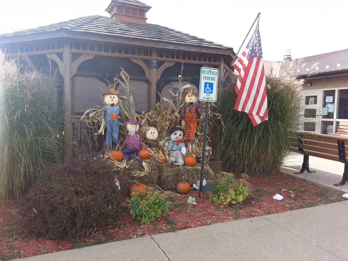 Outdoor entrance area with a wooden gazebo, seasonal scarecrow and pumpkin displays, an American flag, and a handicapped reserved parking sign.