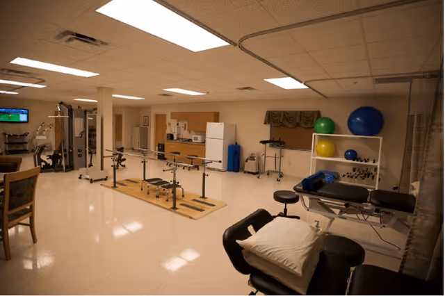 Interior view of a rehabilitation room with exercise equipment including parallel bars, therapy tables, exercise balls, and a television mounted on the wall. The room has a tiled floor, fluorescent ceiling lights, and a kitchenette area with a refrigerator and microwave in the background.