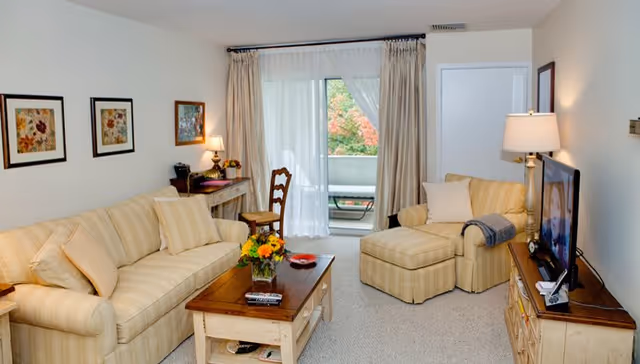 Bright furnished living room with a striped sofa and matching armchair, wooden coffee table, TV stand, desk, and sliding glass door leading to a balcony.