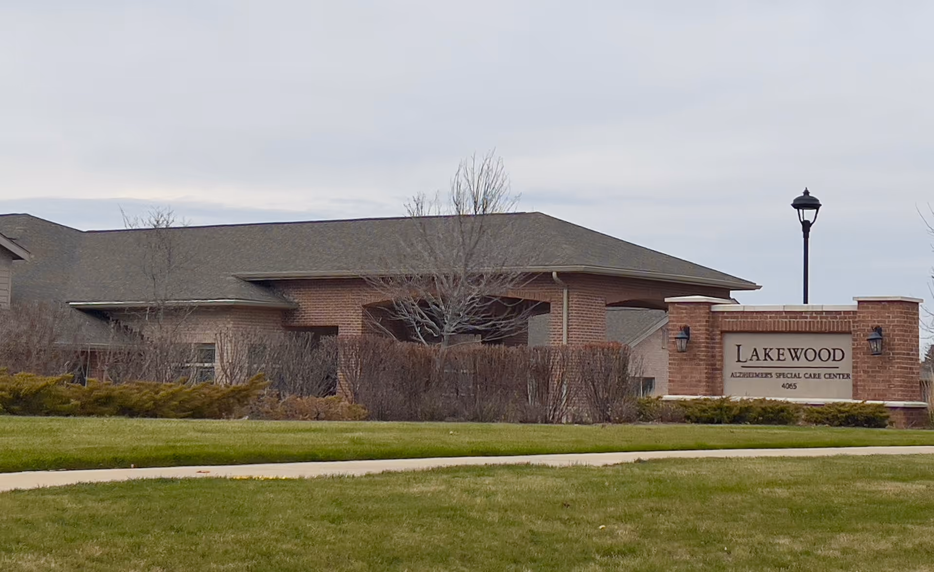 Exterior view of Lakewood Alzheimer's Special Care Center building with a brick sign displaying the facility name. The building has a low-pitched roof and is surrounded by grass, bushes, and a sidewalk. A street lamp is visible near the sign.