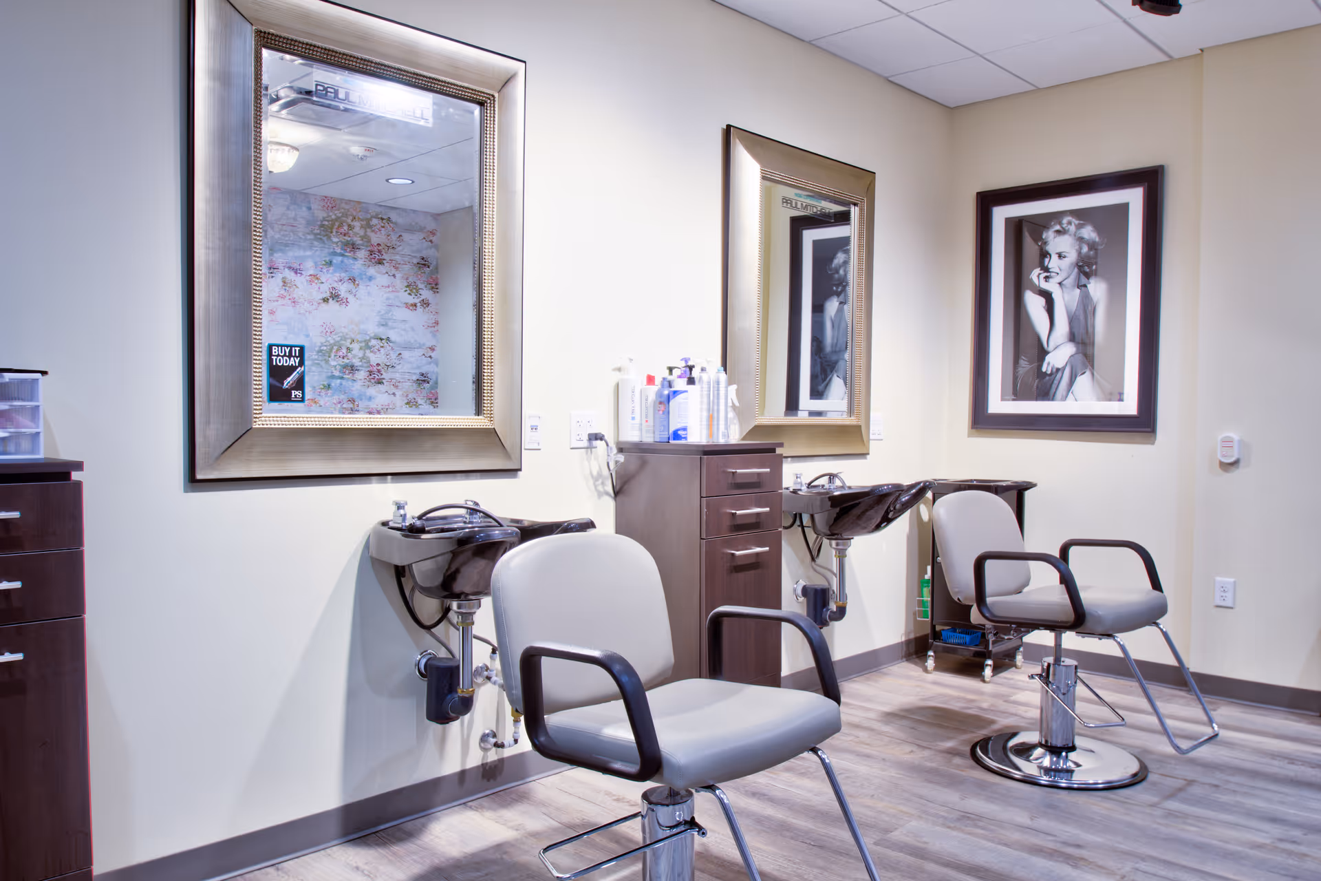 Interior view of a hair salon area with two salon chairs in front of two mirrors mounted on a beige wall. Each station has a black hair washing sink and a small cabinet with hair care products on top. A black and white framed photograph of a woman is hung on the wall to the right.