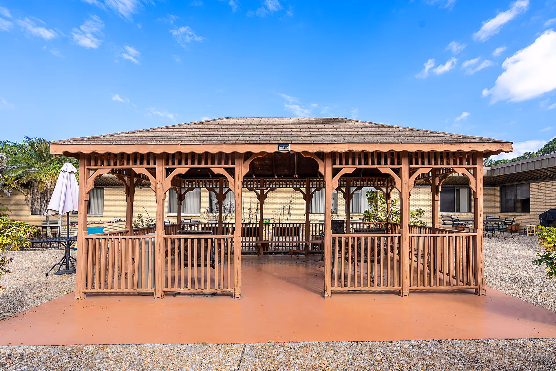 A wooden gazebo with a shingled roof situated in an outdoor courtyard area of a senior living facility. The gazebo has railings and benches inside. Surrounding the gazebo are gravel ground, patio tables with umbrellas, and a beige brick building with windows. The sky is clear with a few clouds.