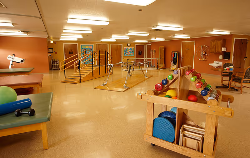 Rehabilitation exercise room with parallel bars, practice stairs, treatment tables and racks of therapy equipment.