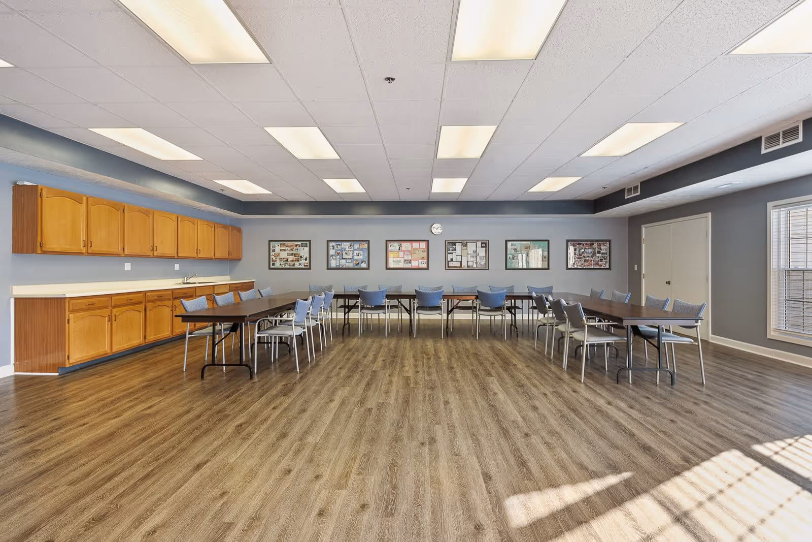 A spacious room with wooden flooring and a row of tables arranged in a U-shape with blue and gray chairs around them. The walls are painted gray with wooden cabinets along one side and several framed pictures and a clock on the opposite wall. Large windows allow natural light to enter the room.