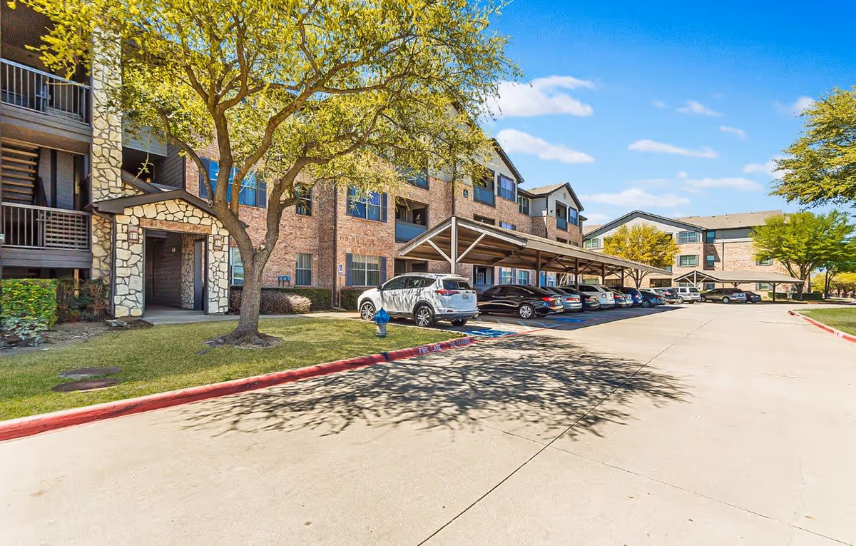 Exterior view of The Cesera 55+ Apartments showing a multi-story brick building with covered parking spaces and several parked cars. There are trees and a grassy area along the sidewalk under a clear blue sky.