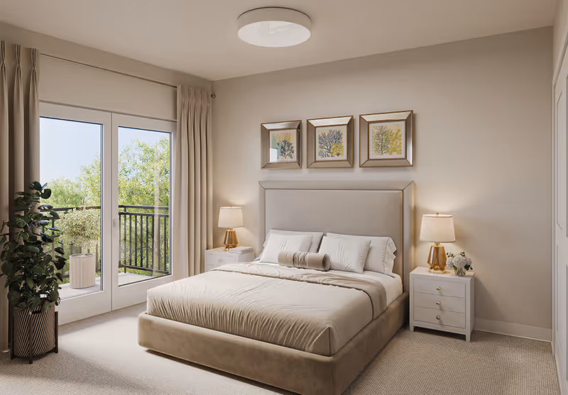 A bright and cozy bedroom featuring a large bed with beige bedding and multiple pillows. On either side of the bed are white nightstands with gold-accented lamps and decorative items. Above the headboard are three framed botanical prints. To the left, there are glass doors leading to a balcony with greenery visible outside. A potted plant is placed near the doors, and beige curtains frame the windows.