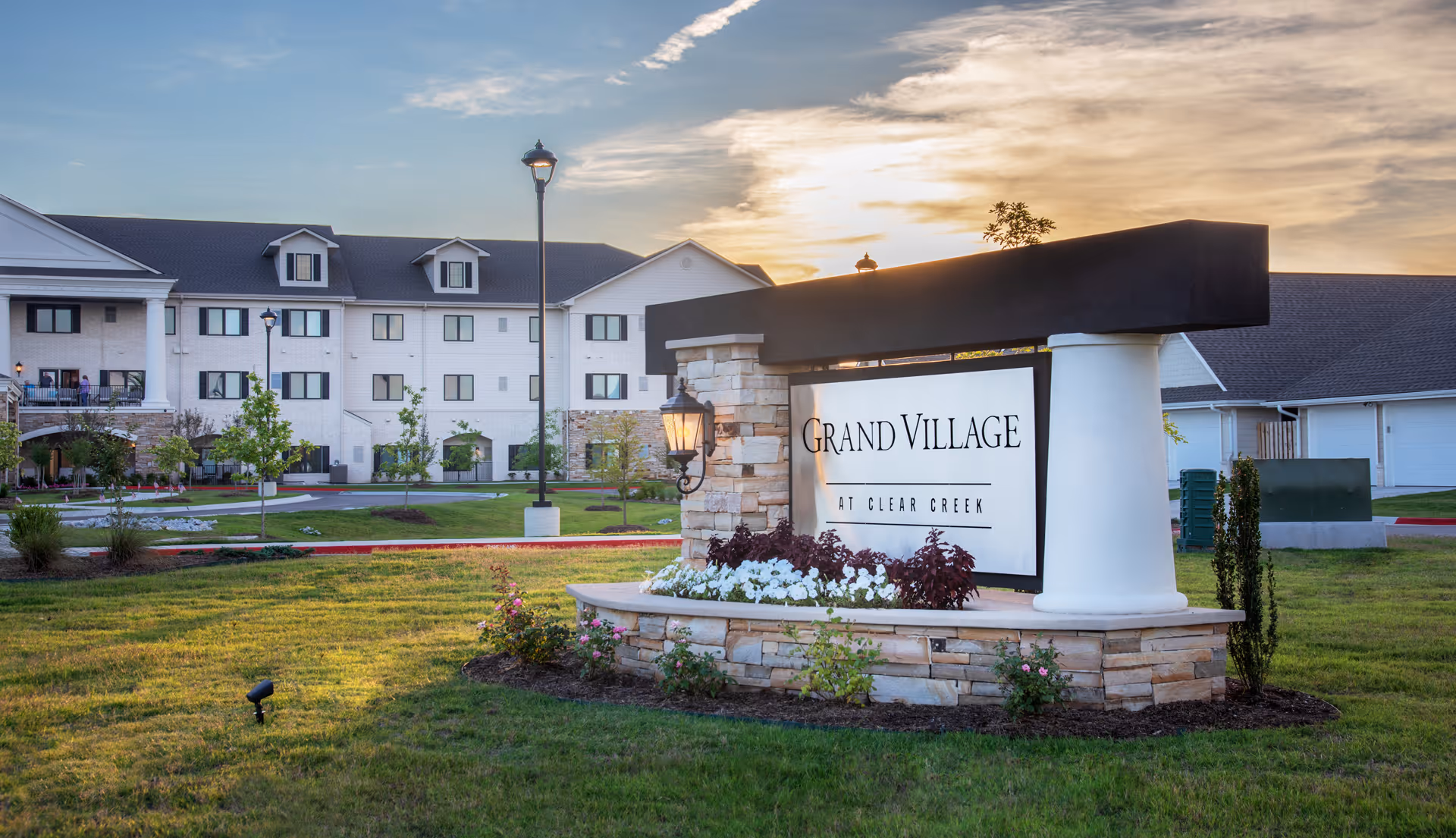 Exterior view of the Grand Village at Clear Creek senior living facility during sunset, showing a large sign with the facility's name surrounded by flowers and greenery, with the building and parking area in the background.