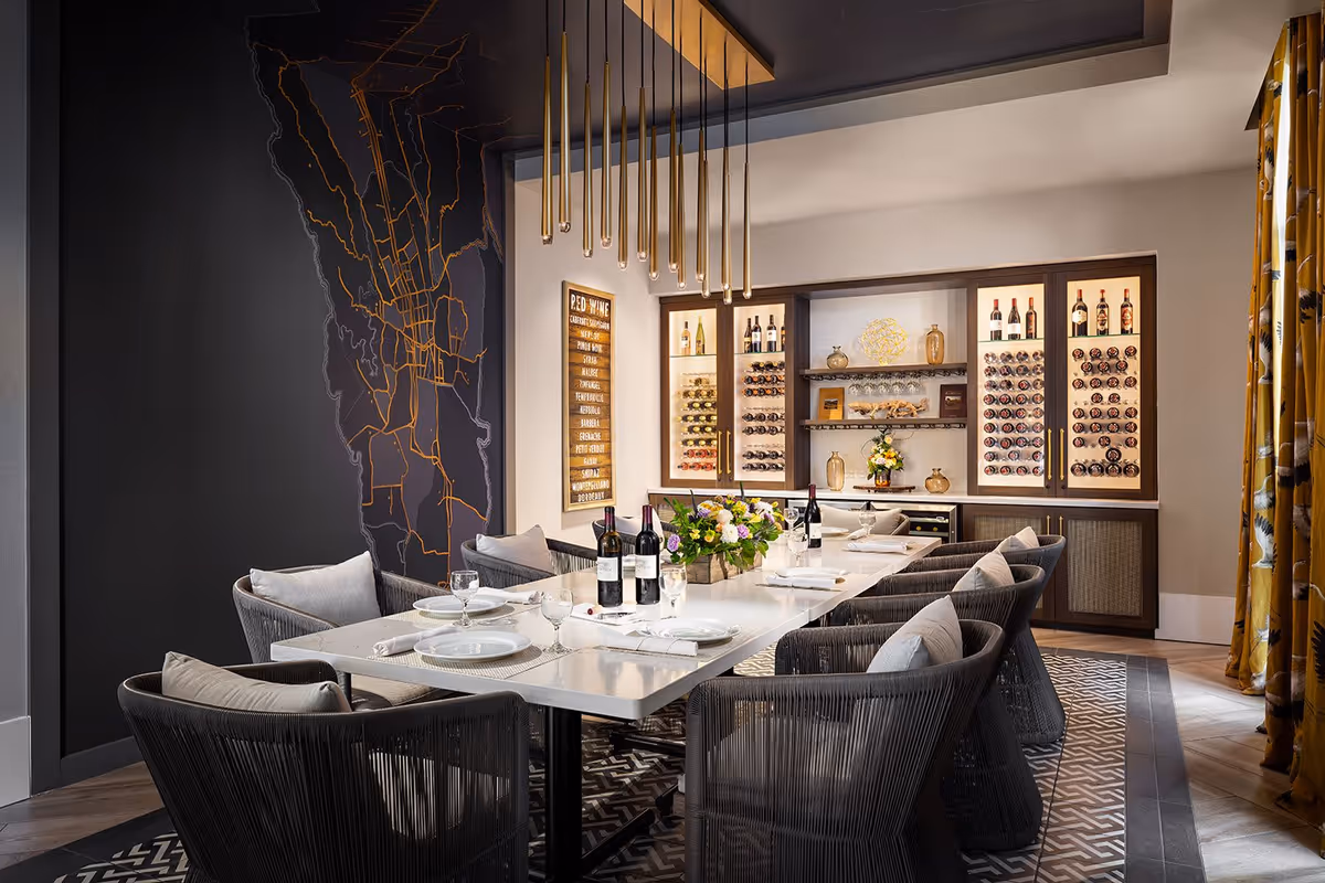A modern dining room with a long white table set for a meal, surrounded by cushioned chairs. The back wall features illuminated wine racks filled with bottles, decorative vases, and a floral centerpiece on the table. A dark wall on the left has a map design in orange lines, and a contemporary gold pendant light fixture hangs above the table. Yellow patterned curtains are visible on the right side.