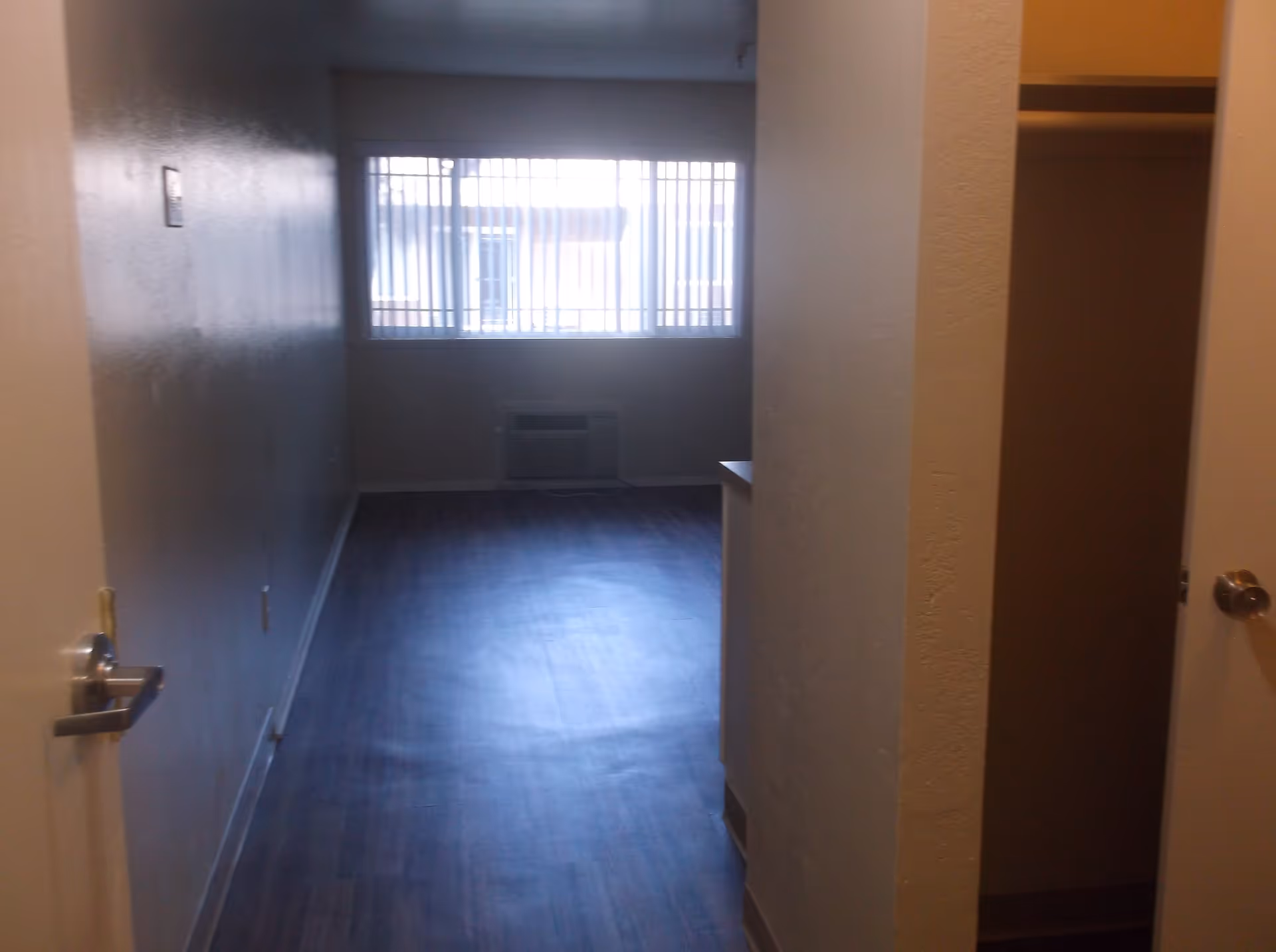 Empty room with dark wood flooring, a window with vertical blinds, an air conditioning unit below the window, and an open closet on the right side.