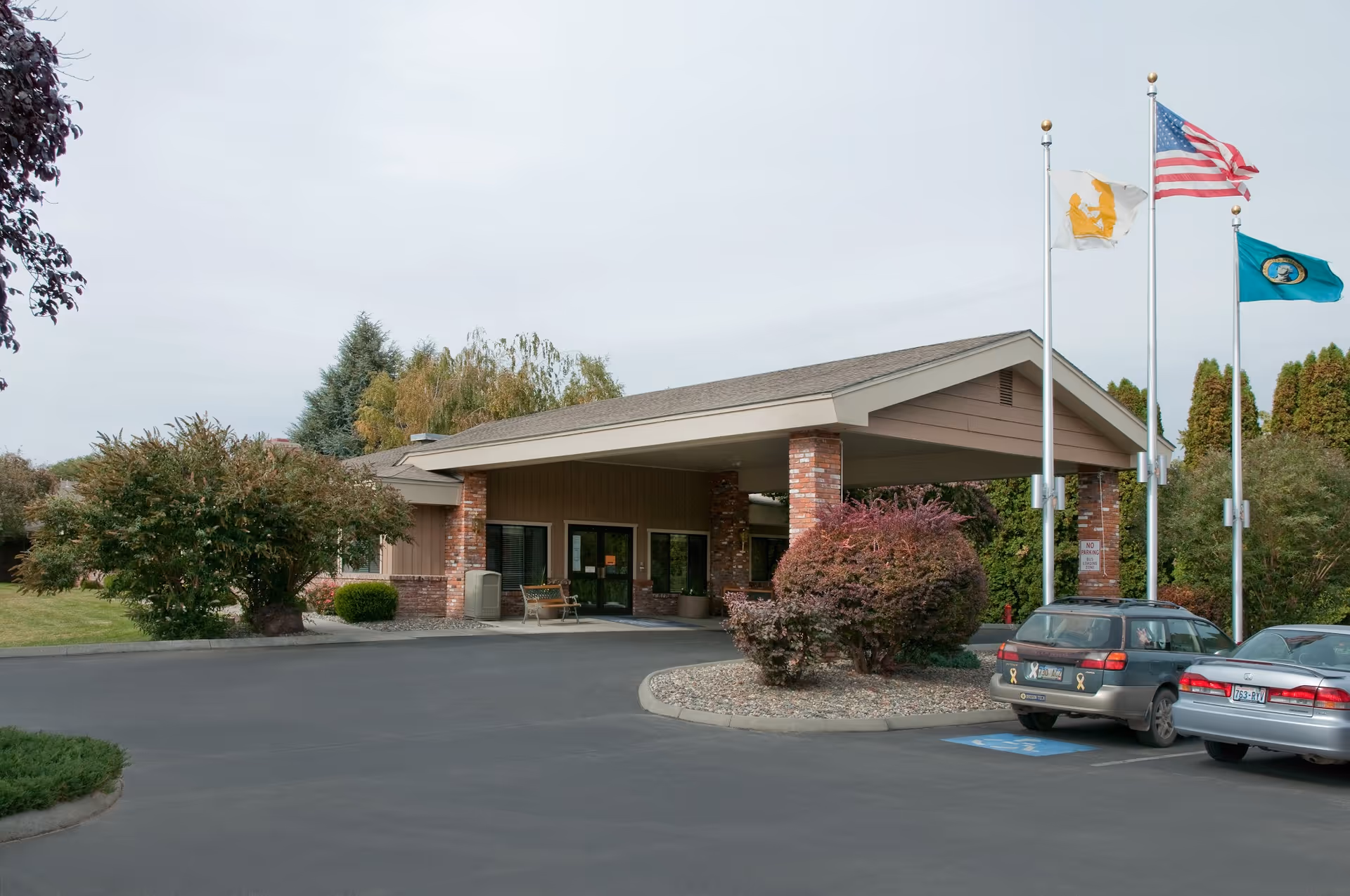Covered entrance and drop-off area of a senior living facility with flagpoles, landscaping, and parked cars.