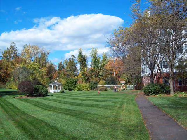 A well-maintained outdoor lawn area with striped grass, a small white gazebo, trees with autumn foliage, a paved walkway on the right, and part of a building visible on the far right under a partly cloudy blue sky.