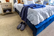 Cozy bedroom with a made bed, nightstand, and slippers on a carpeted floor.