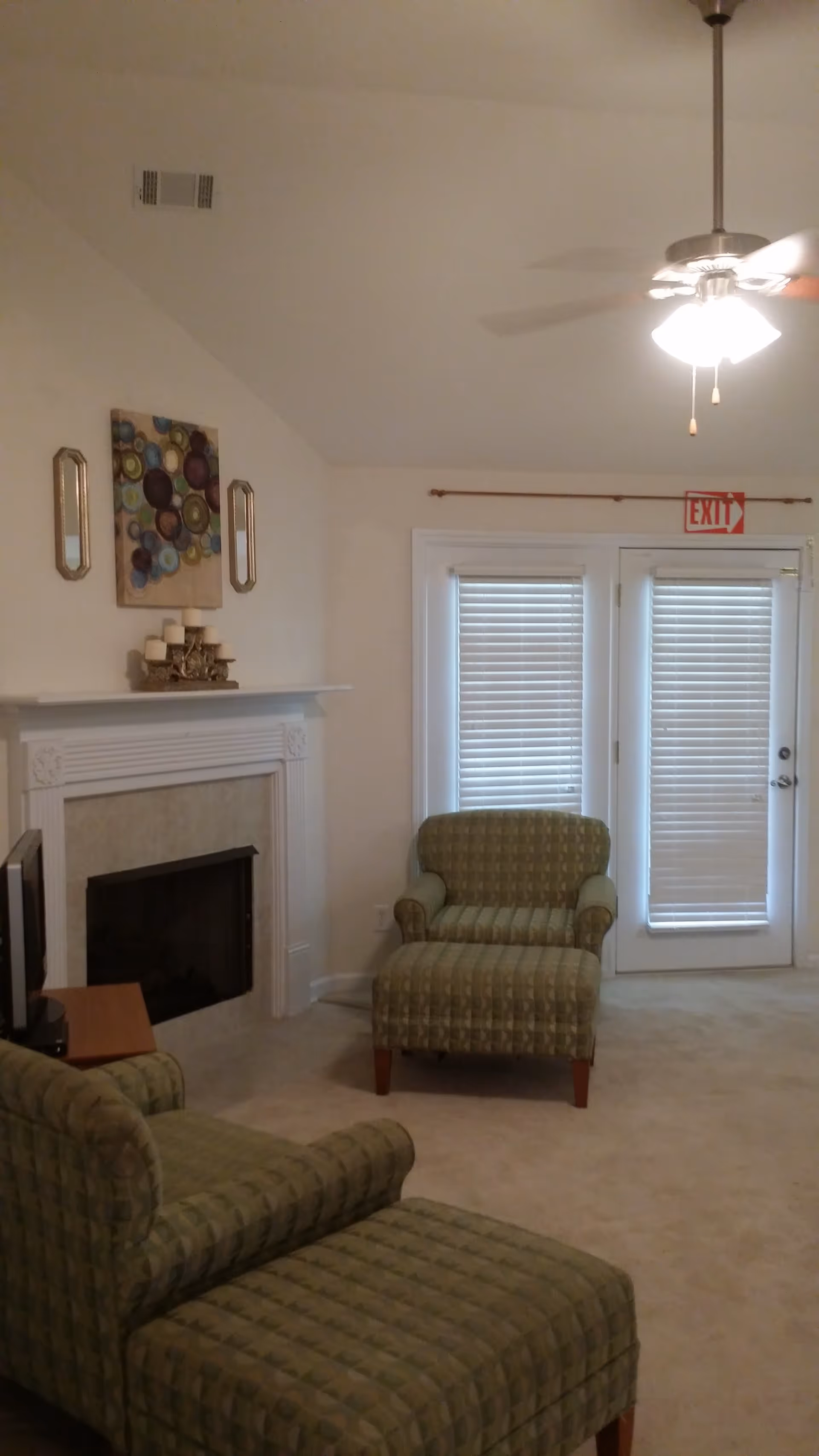 A cozy living room with two green patterned armchairs and matching ottomans, a white fireplace with decorative candles on the mantel, a wall painting with circular designs, a ceiling fan with lights, and a set of French doors with closed blinds and an exit sign above.