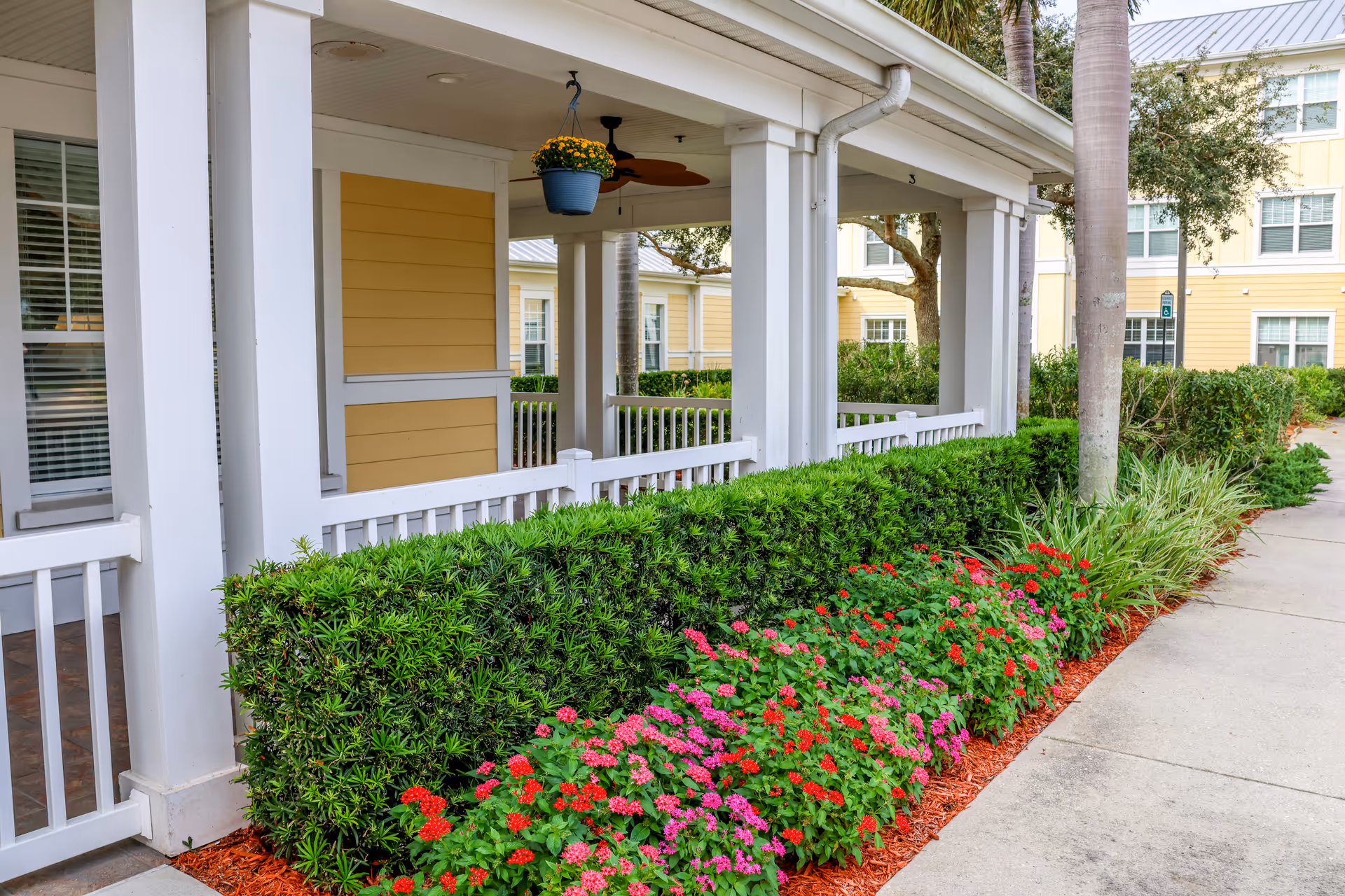 Covered white-columned porch and landscaped walkway with flowering shrubs in front of a yellow senior living building.