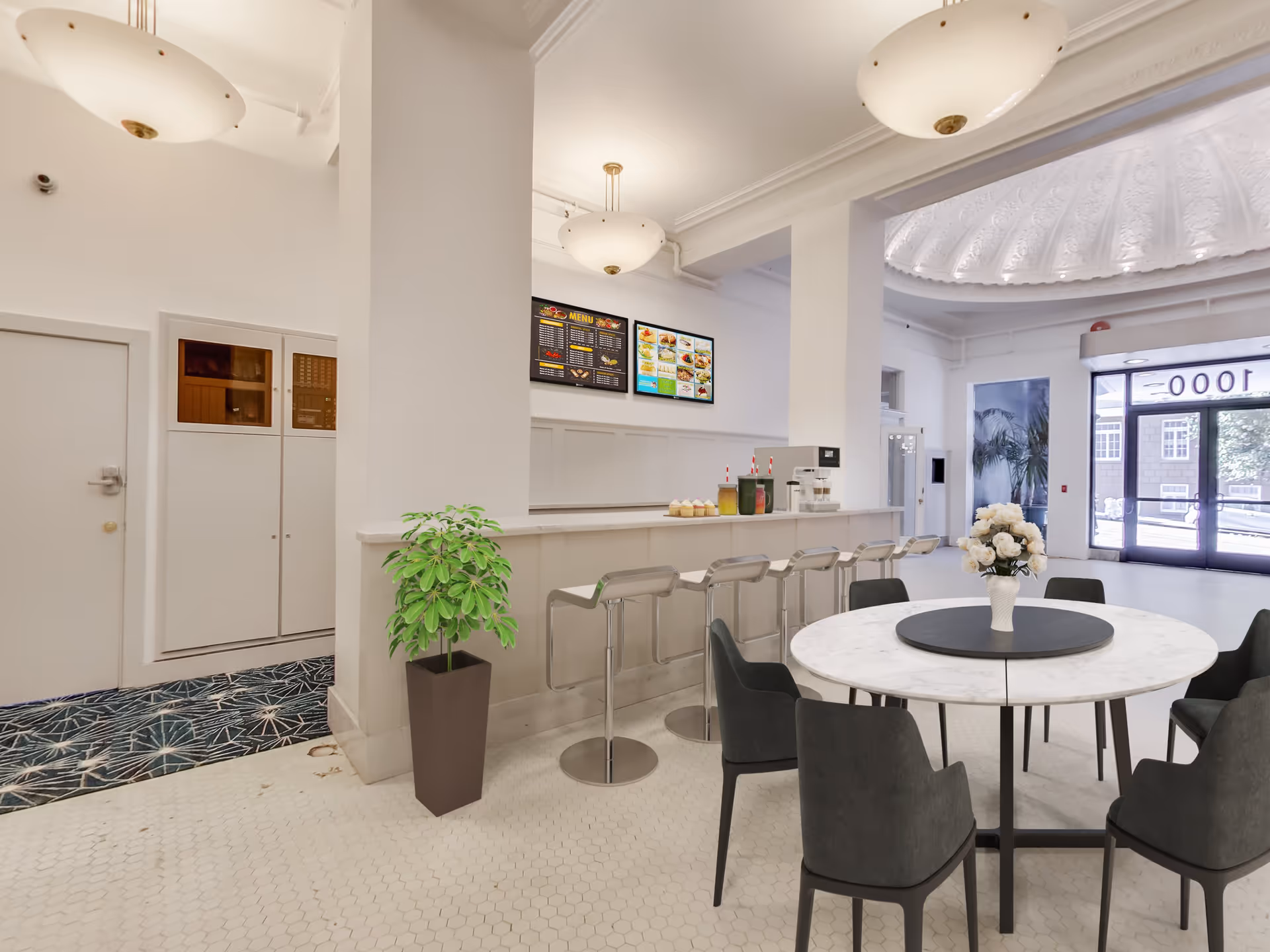 Bright and modern dining area with a round marble table surrounded by six dark chairs, a vase with white flowers on the table, a counter with five bar stools, drinks, and cupcakes. The space features white walls, hexagonal floor tiles, large pendant lights, a potted plant, digital menu boards on the wall, and large glass doors leading outside.