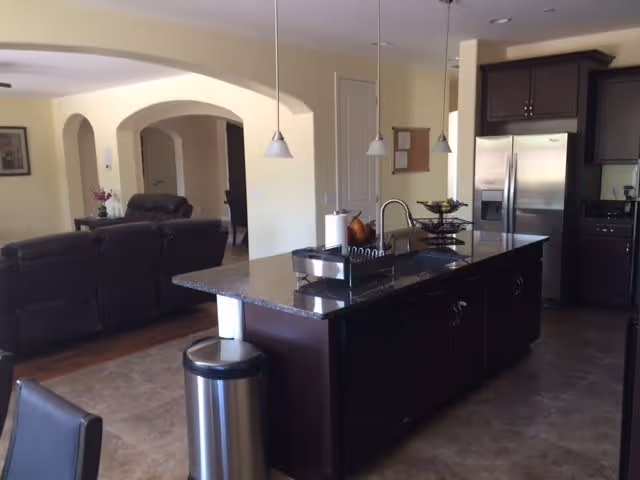 Interior view of a modern kitchen and living room area. The kitchen features a large dark granite island with a sink, paper towel holder, and a tiered fruit basket. Stainless steel refrigerator and dark wood cabinets are visible. The living room has dark leather sofas and is separated by an archway with cream-colored walls.