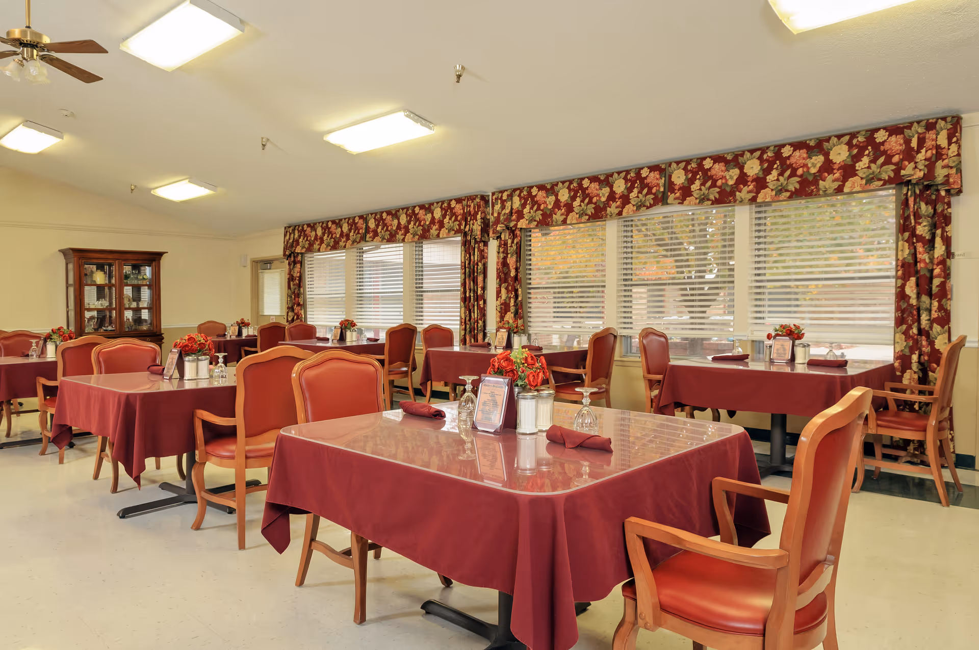 Dining room with multiple tables covered in burgundy tablecloths, each set with napkins, glasses, and small flower arrangements. The room has large windows with floral valances and wooden chairs with red cushions. A wooden cabinet is visible in the background.