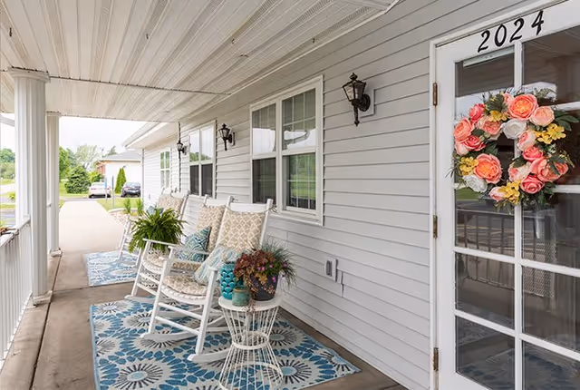Covered porch area at Elkhart Place with white rocking chairs, patterned blue and white rugs, potted plants, and a door with the number 2024 and a colorful floral wreath.