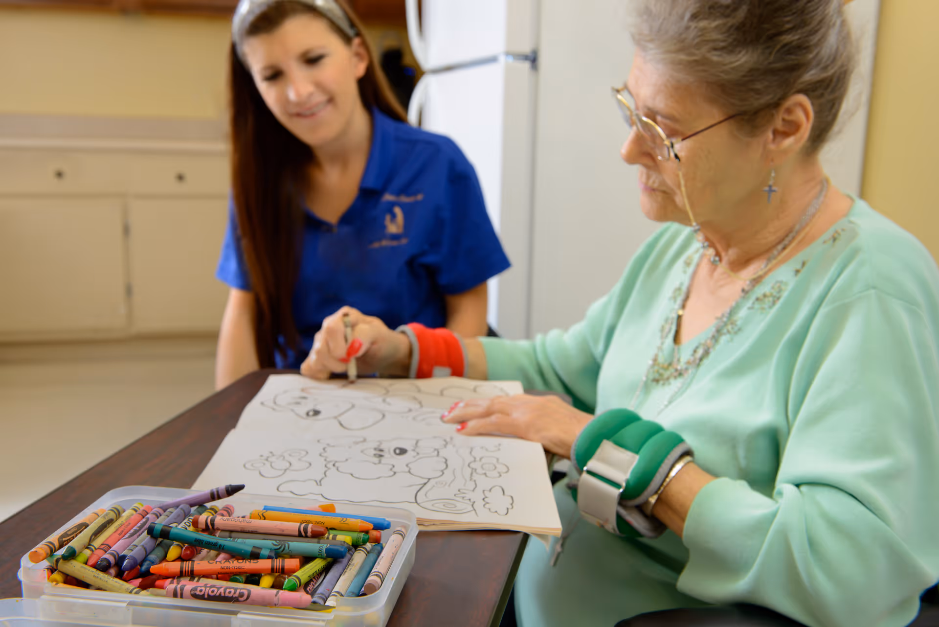 An elderly woman with wrist weights is coloring a picture in a coloring book at a table, while a younger woman in a blue shirt watches and smiles. A container of crayons is on the table in front of them.