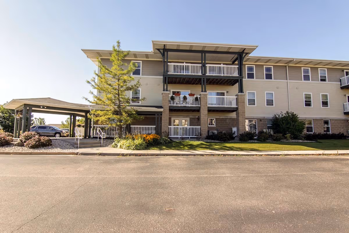 Exterior view of a three-story senior apartment building with balconies, a covered entrance, landscaping with bushes and a tree, and a parked car nearby under a clear sky.