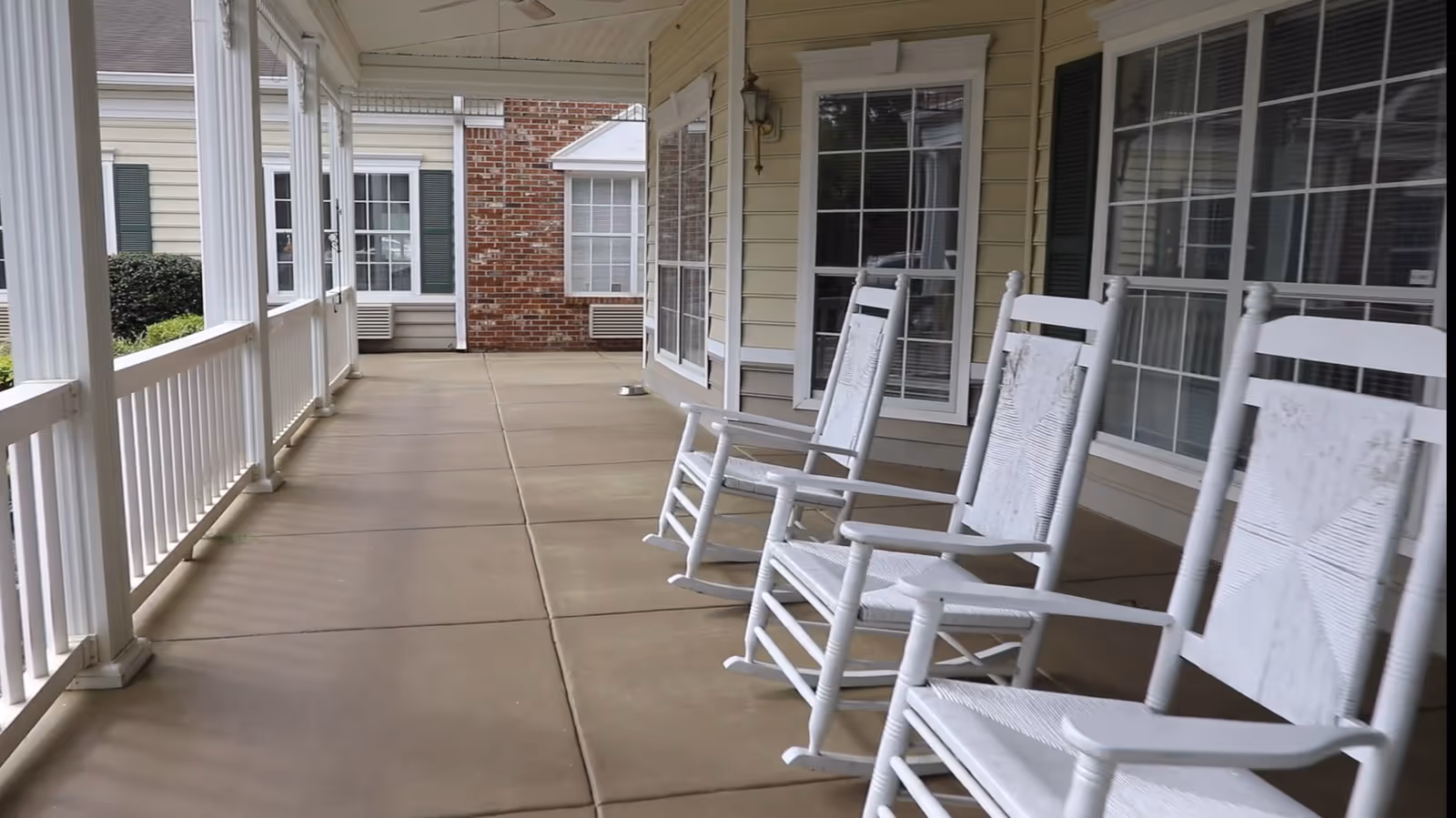 A covered outdoor porch area with three white wooden rocking chairs lined up along the wall of a building. The porch has a beige tiled floor, white railings, and large windows with green shutters. The building exterior features beige siding and some brickwork.