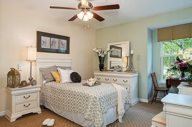 A cozy bedroom with a single bed covered in a patterned gray and white bedspread, several decorative pillows, and a tray with a teapot and cup. The room features a white nightstand with a lamp and decorative items, a white dresser with a large mirror and flower arrangements, a wooden chair by a window with a view of greenery outside, and a ceiling fan with lights.