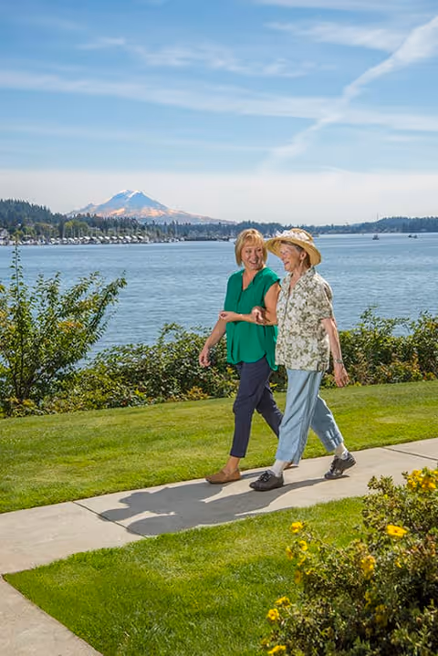 Two women walking arm in arm on a paved path beside a grassy area with bushes and yellow flowers, overlooking a large body of water with a mountain in the background under a clear blue sky.
