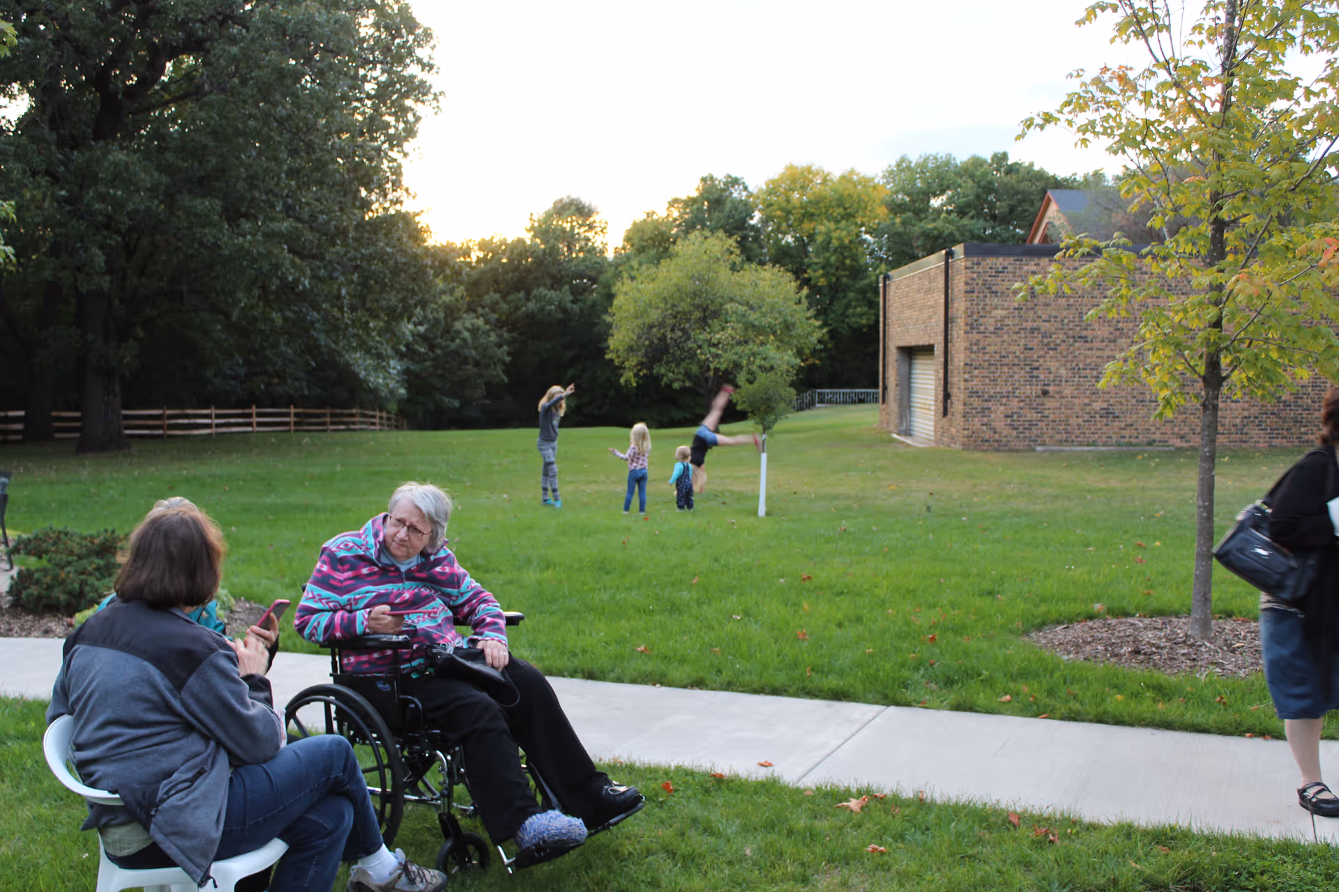 An elderly woman in a wheelchair and another woman sitting on a chair are conversing outdoors on a grassy area with a sidewalk. In the background, several children are playing on the grass near a brick building surrounded by trees.