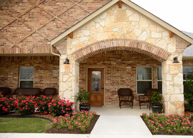 Front entrance of a brick and stone building with a covered porch featuring two wicker chairs and potted plants. There are flower beds with pink flowers on either side of the concrete walkway leading to a wooden door with decorative glass.
