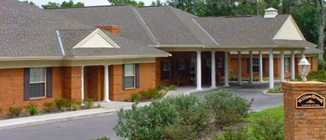 Exterior view of WillowBrook Assisted Living facility showing a single-story brick building with a covered entrance supported by white columns, surrounded by greenery and a paved driveway.