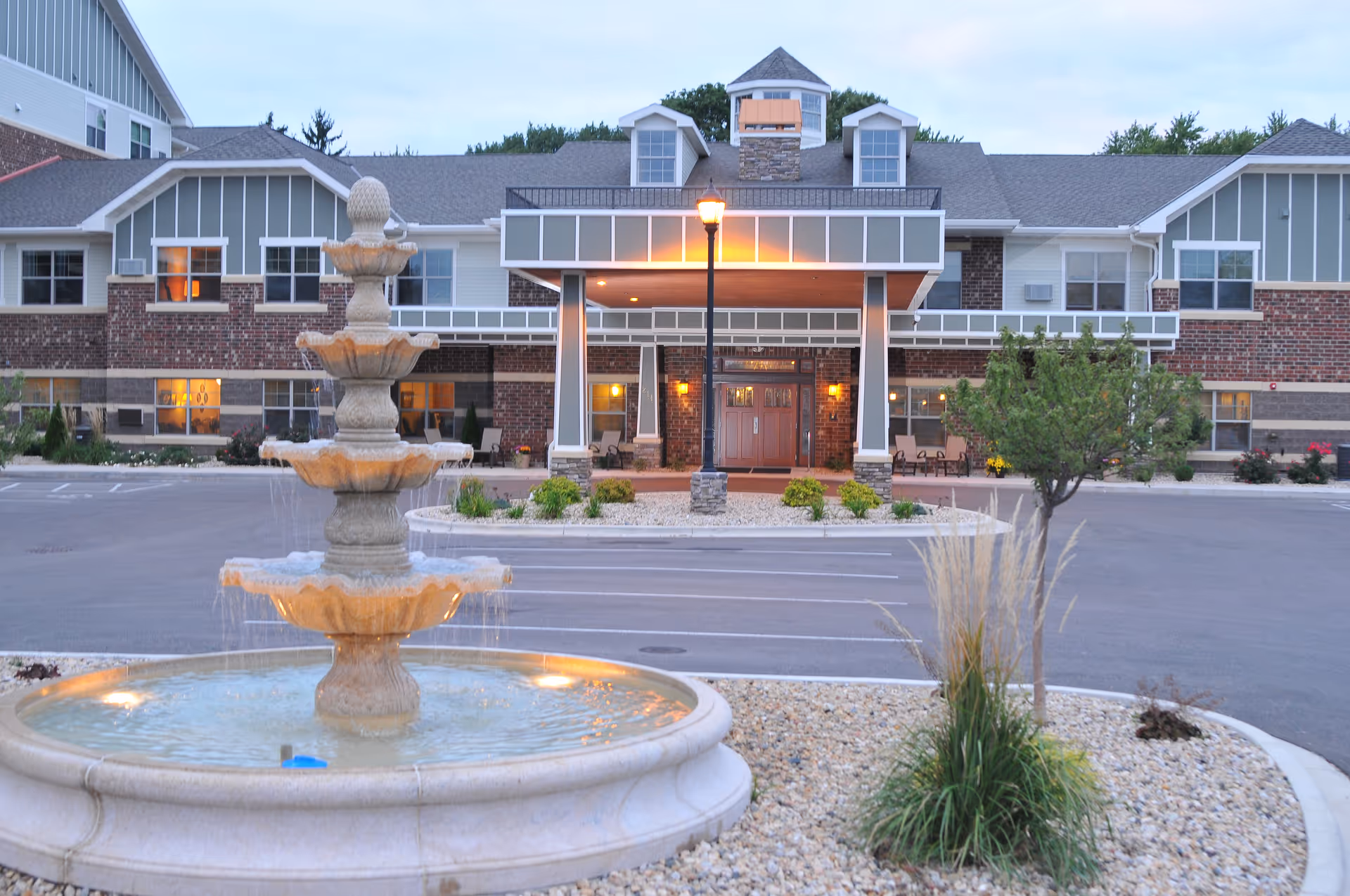 Front exterior view of Heritage Senior Campus building at dusk with a multi-tiered stone fountain in the foreground, a lamp post, landscaped rocks, small trees, and shrubs surrounding the entrance.