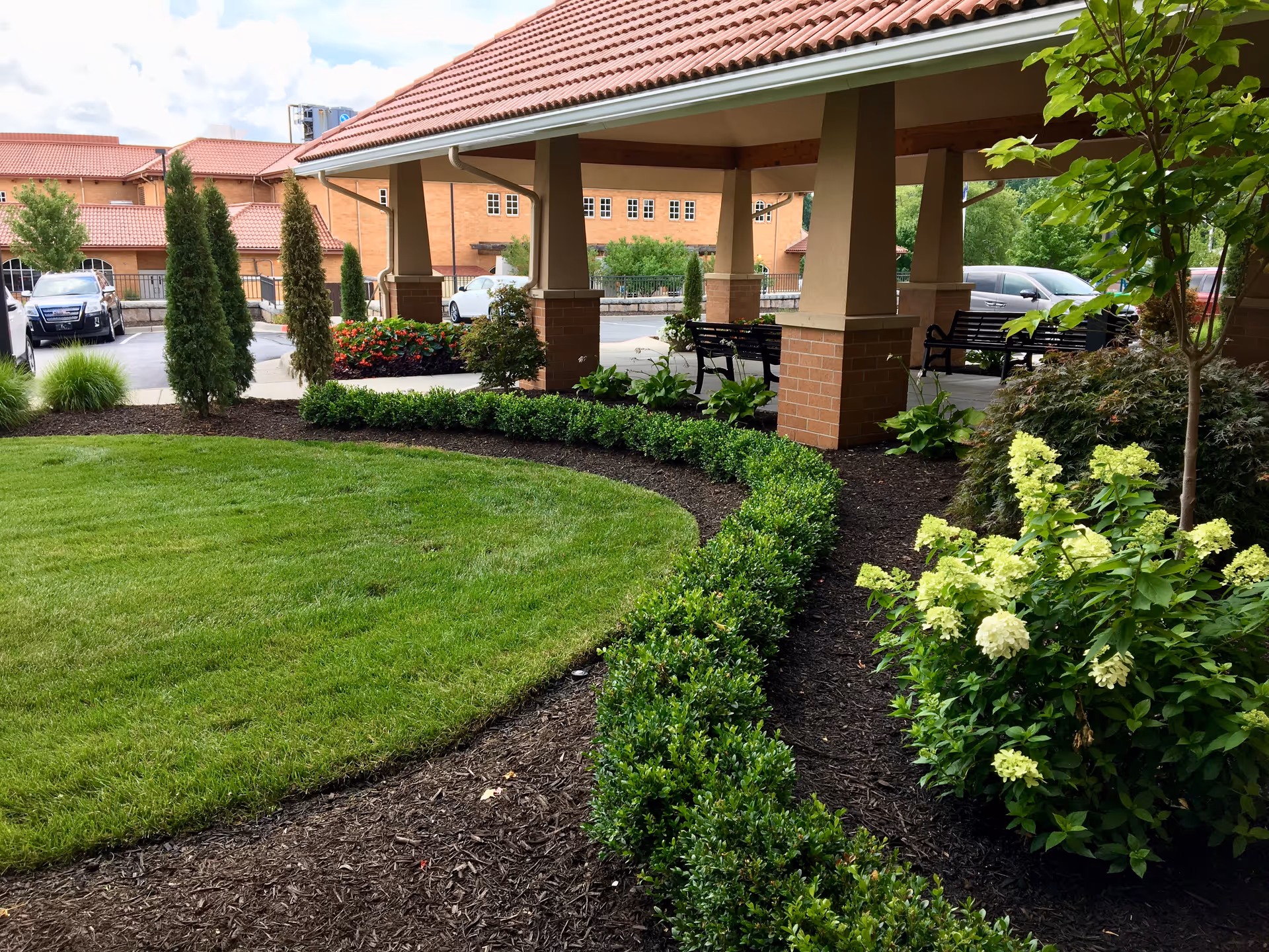 A landscaped outdoor area at Mission Square Independent Senior Living featuring a well-maintained lawn, neatly trimmed bushes, flowering plants, and a covered seating area with benches. In the background, there are parked cars and a building with a red-tiled roof.